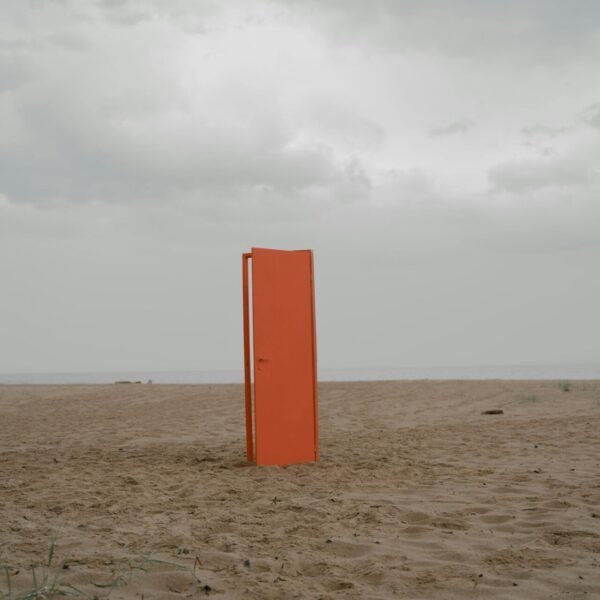 An orange door stands alone on an empty beach under a cloudy sky.