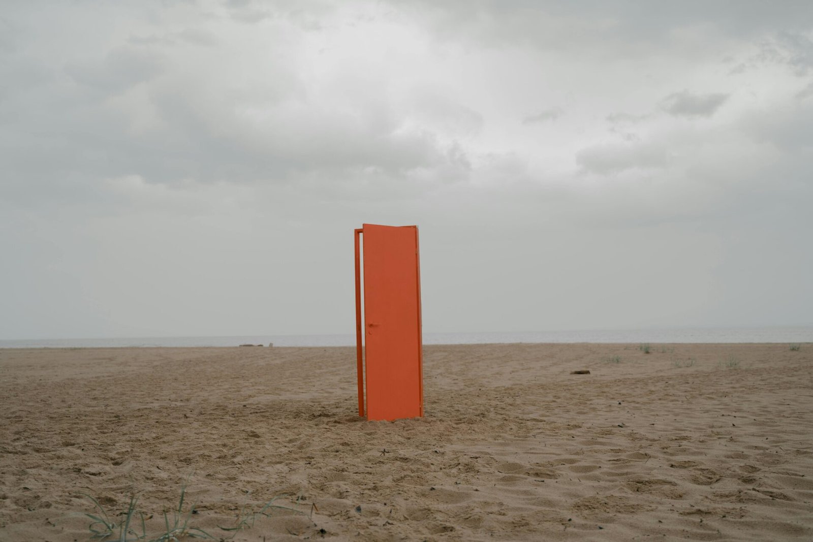 An orange door stands alone on an empty beach under a cloudy sky.